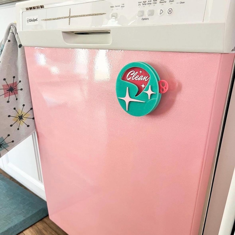 May include: A pink dishwasher with a turquoise and red "Clean" magnet attached to the door. The magnet has a starburst design and a small red circle with a white dot.