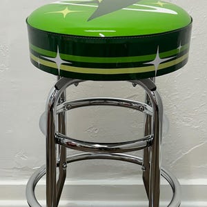 Puede incluir: Un taburete de bar verde y blanco con base cromada. El asiento est&aacute; tapizado en vinilo verde con un dise&ntilde;o de estrella blanca.