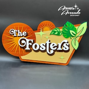 May include: A retro-style sign with the name "The Fosters" in white, set against a warm color palette of orange, yellow, and brown. The design includes a sunburst, starbursts, and green leaf accents, with the "Atomic Avocado Designs" logo.