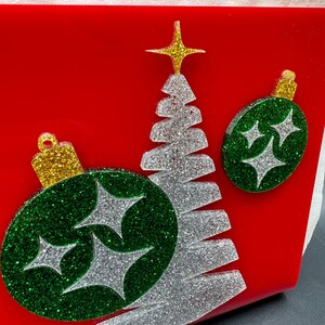 May include: A red background with a silver glitter Christmas tree and two green glitter ornaments with silver glitter stars. The ornaments have a gold glitter top.