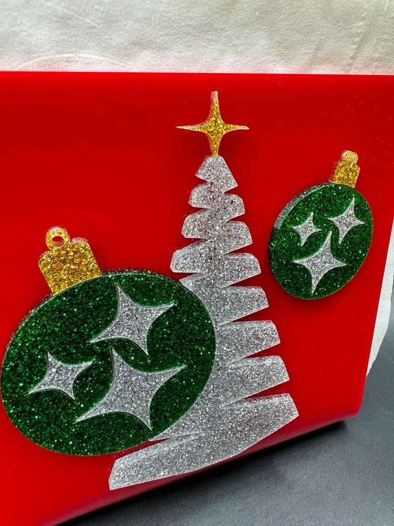 May include: A red background with a silver glitter Christmas tree and two green glitter ornaments with silver glitter stars. The ornaments have a gold glitter top.