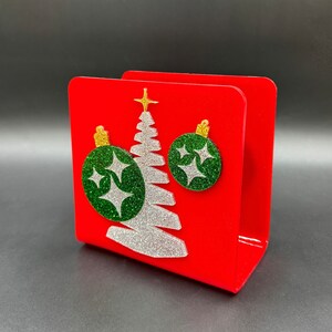 May include: Red napkin holder with a white glitter Christmas tree and green glitter ornaments. The ornaments have white glitter stars on them.