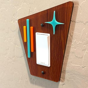 May include: A light switch cover with a mid-century modern design. The cover is made of wood and features a teal star, orange and teal stripes, and a white light switch. The text "Insteon" is visible on the switch.