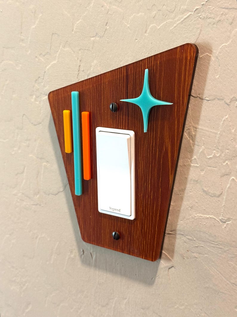 May include: A light switch cover with a mid-century modern design. The cover is made of wood and features a teal star, orange and teal stripes, and a white light switch. The text "Insteon" is visible on the switch.