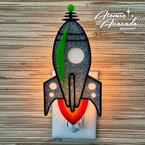 May include: A silver glitter rocket ship night light with a green fin and orange flames. The light plugs into a standard outlet.  Atomic Avocado Designs is printed on the image.