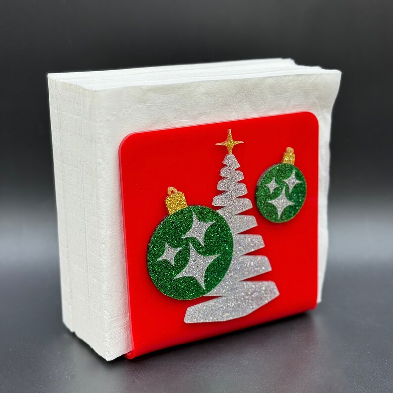 May include: A red and white Christmas-themed napkin holder with a glitter-covered tree and ornaments. The holder is made of wood and has a white napkin inside.
