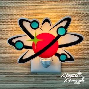 May include: A white night light with a retro atomic design. The design features a red sphere, black orbiting lines, teal circles, and a green star. The night light has the text "Atomic Avocado DESIGNS" in a script font.