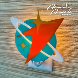 May include: A white night light shaped like a planet with a red and orange star shooting through it. The planet is turquoise blue with a white ring around it. The night light is plugged into an outlet.
