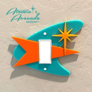 以下が含まれることがあります： ターコイズ、オレンジ、イエローのミッドセンチュリーモダンなライトスイッチカバーで、スターバーストのデザインが施されています。  Atomic Avocado Designs®がカバーの上部に印刷されています。