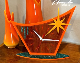 May include: Orange and wood clock with a starburst design and a green glitter base. The clock face has a wood grain pattern and white hands. The text "Atomic Avocado Designs" is visible in the top right corner of the image.