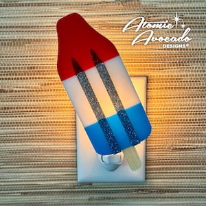 May include: A red, white, and blue popsicle-shaped night light with glitter accents and wooden sticks. The night light is plugged into a white outlet. The text "Atomic Avocado Designs" is in the upper right corner.