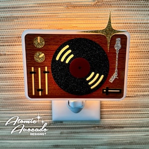 May include: A retro-style night light shaped like a record player. The design features a brown wood-grain background, a black vinyl record, and a white frame. Accents include gold glitter and the text "Atomic Avocado Designs®".