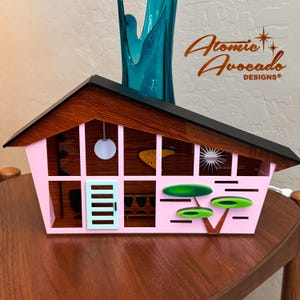 Puede incluir: Una casa en miniatura de color rosa y marrón con un techo negro, que presenta una puerta azul claro y formas geométricas verdes. La casa está iluminada desde el interior y tiene una bombilla blanca colgando del techo. El texto "Atomic Avocado Designs" es visible en la esquina superior derecha de la imagen.