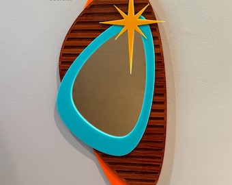 May include: A mid-century modern style wall mirror with a teal blue frame, orange and brown accents, and a gold starburst design.  Atomic Avocado Designs is printed on the top left corner.