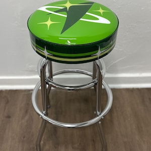 Puede incluir: Un taburete de bar verde con estructura cromada. El asiento es redondo y presenta un dise&ntilde;o retro con una nave espacial blanca y estrellas.