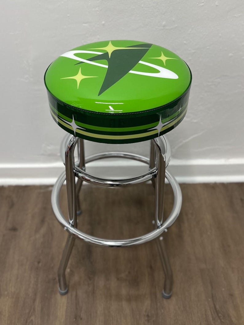 Puede incluir: Un taburete de bar verde con estructura cromada. El asiento es redondo y presenta un dise&ntilde;o retro con una nave espacial blanca y estrellas.