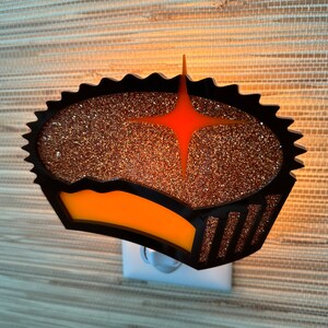 May include: A black and orange nightlight shaped like a cupcake with a star on top. The cupcake is covered in glitter.