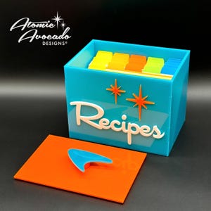 Puede incluir: Una caja de recetas azul turquesa con una etiqueta blanca que dice "Recetas" y dos estrellas naranjas. La caja tiene una tapa naranja extraíble con un asa azul en forma de átomo.