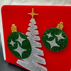 May include: A red acrylic napkin holder with a silver glitter Christmas tree and two green glitter ornaments with silver glitter stars.