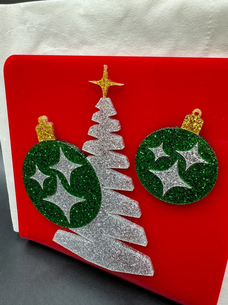May include: A red acrylic napkin holder with a silver glitter Christmas tree and two green glitter ornaments with silver glitter stars.