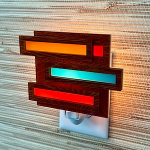 May include: A wooden night light with a geometric design featuring orange, red, and blue lights. The night light plugs into a standard electrical outlet.