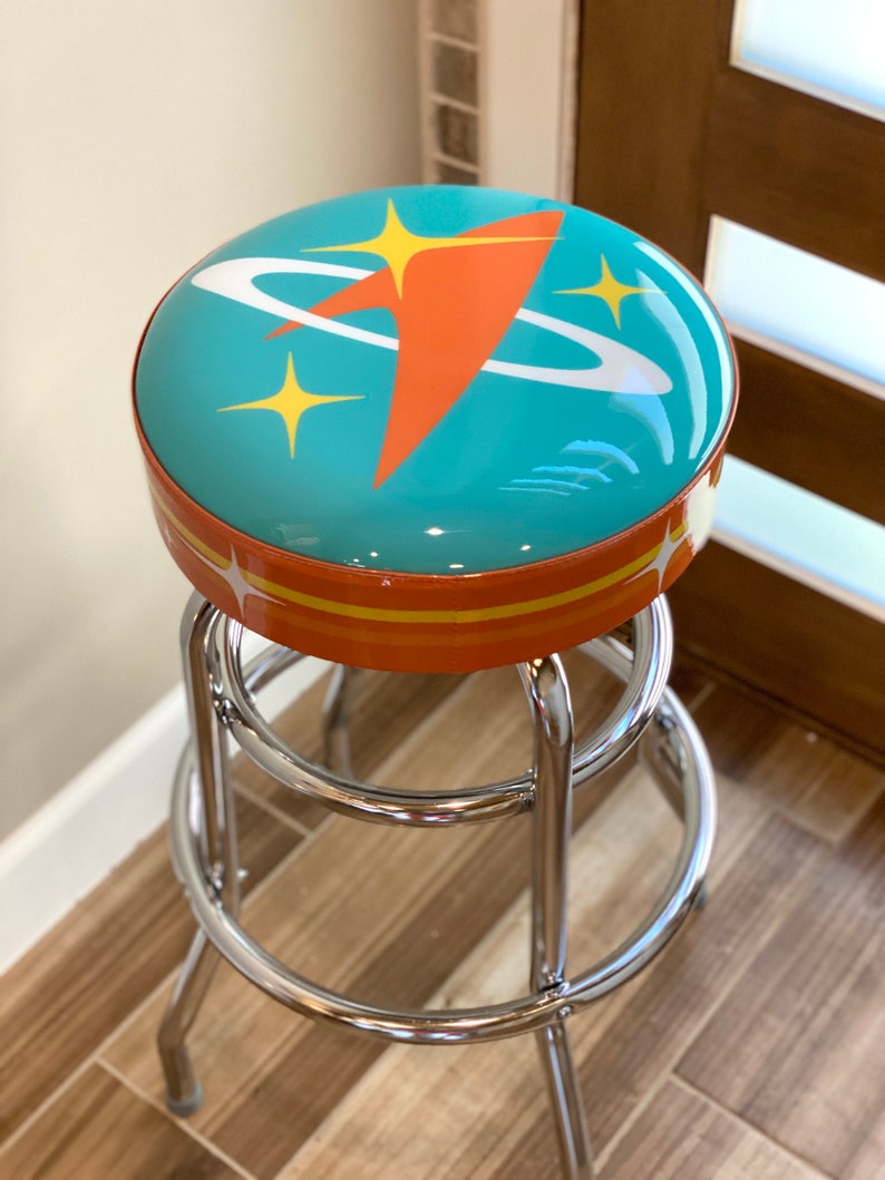 Puede incluir: Un taburete de bar turquesa y naranja con un dise&ntilde;o retro de tem&aacute;tica espacial. El asiento presenta una nave espacial estilizada con un anillo blanco a su alrededor y estrellas amarillas.