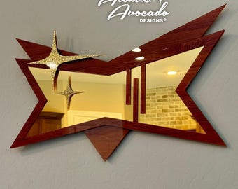 May include: A wall mirror with a dark wood grain frame in a sharp, angular starburst shape. The mirror features two gold glitter starburst accents on the left side and three vertical rectangular slats on the right side. The text Atomic Avocado DESIGNS is visible.