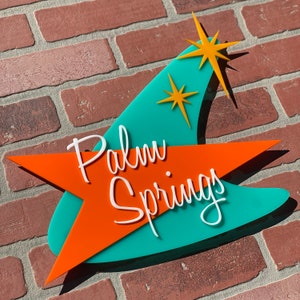 K&ouml;nnte beinhalten: Ein retro-m&auml;&szlig;iges Schild in T&uuml;rkis und Orange mit einem Sternburst-Design und dem Text "Palm Springs".
