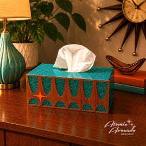 May include: A rectangular tissue box with a teal glitter top and a wooden front featuring an orange starburst design. White tissues are pulled from the top. The Atomic Avocado Designs logo is visible.