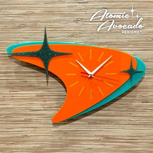 May include: Orange and teal mid-century modern clock with a boomerang shape and glitter stars. The clock face has white hands and gold hour markers. The text "Atomic Avocado Designs" is in the top right corner.