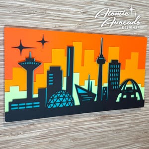 May include: A layered wall art piece featuring a stylized cityscape silhouette in black against a gradient background of orange, yellow, and light green. The artwork includes the text "Atomic Avocado Designs".