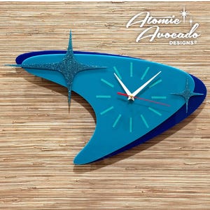May include: A retro-style wall clock in shades of blue, shaped like a boomerang. The clock features a turquoise face with white hands and a red second hand. Two glittery starbursts and the text "Atomic Avocado Designs" add to the mid-century modern aesthetic.
