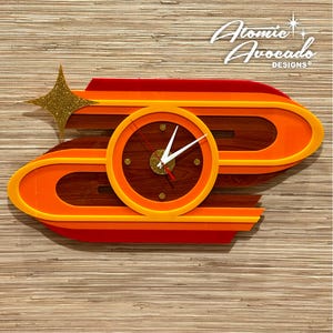 May include: A retro-style wall clock with a mid-century modern design. The clock features a circular face with white hands and a gold-colored center, set against a dark wood grain background. The clock is surrounded by orange and red layered shapes, with a gold star accent.