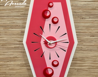 May include: A pink and red atomic-style clock with a white frame and black clock hands. The clock face has a white center circle with a red ring around it. The clock is surrounded by a pink and red background with a white border.