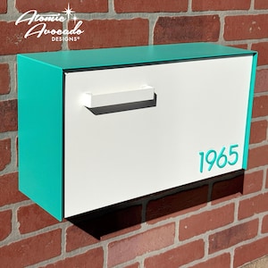 May include: A modern mailbox with a white front and a teal top and sides. The number "1965" is displayed in teal on the front. The mailbox is mounted on a red brick wall. The brand name "Atomic Avocado Designs" is in the upper left corner.