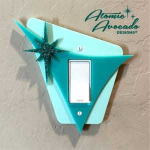 Puede incluir: Una cubierta de interruptor de luz de mediados de siglo moderna en color turquesa y azul claro con un acento de estrella brillante. La cubierta tiene un diseño triangular y el texto "Atomic Avocado DESIGNS®".