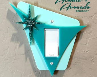 May include: A teal and light blue mid-century modern light switch cover with a glittery starburst accent. The cover has a triangular design and the text "Atomic Avocado DESIGNS®".