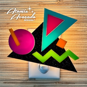 Puede incluir: Una luz nocturna con forma de diseño geométrico con fondo negro, formas verdes, azules, naranjas y rosas. La luz nocturna está enchufada a una toma de corriente blanca. El texto "Atomic Avocado Designs" es visible en la esquina superior izquierda de la imagen.