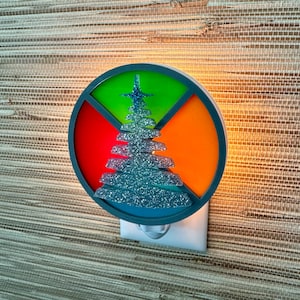 Può includere: Un notturno rotondo e colorato con un design verde, rosso, arancione e blu. Il centro del notturno presenta un albero di Natale con glitter argentati.