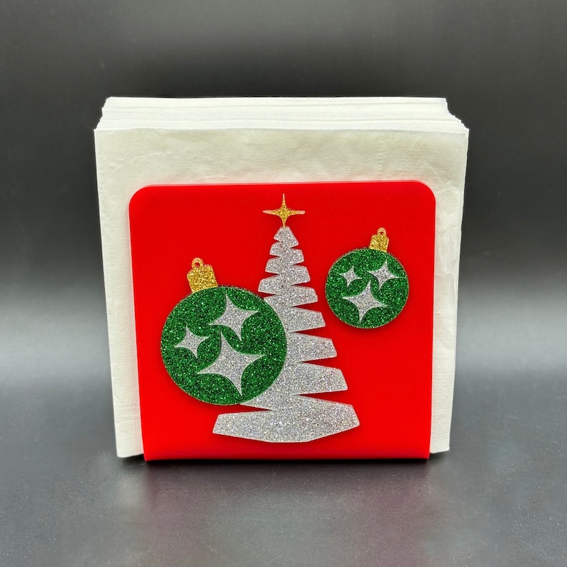 May include: A red napkin holder with a white napkin and a silver glitter Christmas tree with green glitter ornaments. The tree has a gold star on top.