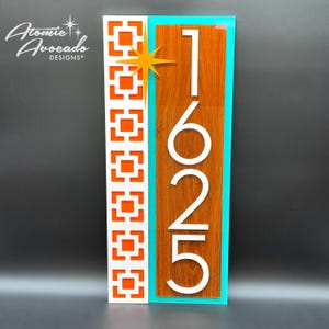 Puede incluir: Un letrero de número de casa moderno con fondo blanco y patrón geométrico naranja. El número de casa 1625 se muestra en blanco sobre un fondo de madera con borde turquesa.