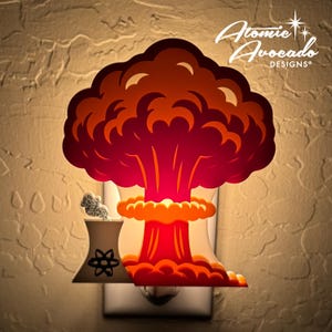 Puede incluir: Una luz nocturna en forma de hongo atómico rojo y naranja con una base blanca y un símbolo de átomo. La luz nocturna está enchufada a una toma de corriente. El texto "Atomic Avocado Designs" es visible en la esquina superior derecha de la imagen.