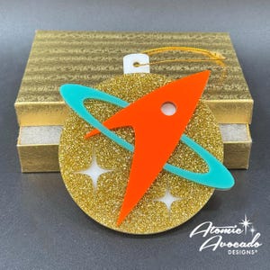 May include: A gold glitter ornament with an orange rocket ship design and a teal ring. The ornament has two white starbursts and a white hanging loop. The text "Atomic Avocado Designs" is in the bottom right corner.