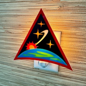 Könnte beinhalten: Ein dreieckiges Nachtlicht mit rotem Rand und schwarzem Hintergrund. Das Nachtlicht zeigt ein stilisiertes Bild der Erde mit einer aufgehenden roten Sonne, einem Kometen und Sternen. Das Nachtlicht ist in eine weiße Steckdose gesteckt.