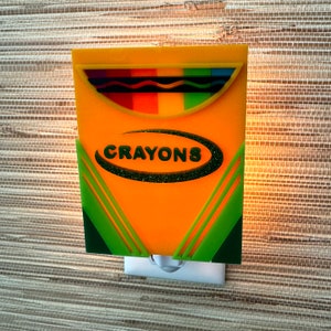 May include: A yellow night light shaped like a box of crayons. The box has a green and white border and the word "CRAYONS" is printed in black letters on the front.