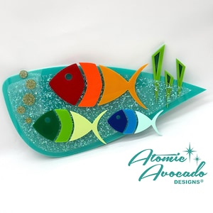 Op de afbeelding: Een turquoise blauwe wanddecoratie van hars met drie gestileerde vissen. De vissen zijn gemaakt van gelaagd acryl in tinten rood, oranje, geel, groen, blauw en wit. De achtergrond is getextureerd met gouden glitter. Het stuk is gesigneerd met "Atomic Avocado Designs".