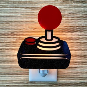 May include: A black and white night light shaped like a vintage video game joystick with a red button on top. The light is plugged into a standard electrical outlet.