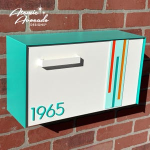 Puede incluir: Un buzón moderno de mediados de siglo con cuerpo blanco, parte superior y laterales turquesa y asa blanca. El buzón presenta rayas verticales en turquesa, naranja y rojo. El número "1965" se muestra en turquesa. Montado en una pared de ladrillo.