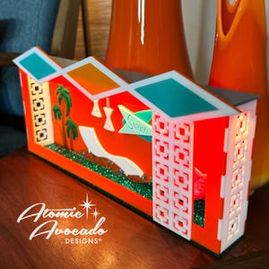 Puede incluir: Una casa de estilo moderno de mediados de siglo, roja y blanca, con una piscina verde y palmeras. La casa tiene un techo blanco con un borde turquesa y una valla blanca. La casa está iluminada con luces rojas. El texto "Swim" está en un letrero verde.