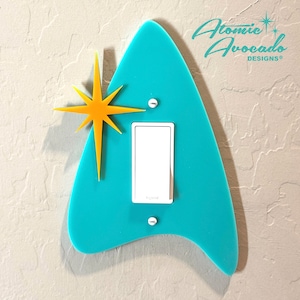 May include: A turquoise blue light switch cover with a yellow starburst design. The cover is shaped like a stylized arrowhead and has a white light switch in the center. The text "Atomic Avocado Designs" is printed in the top right corner.